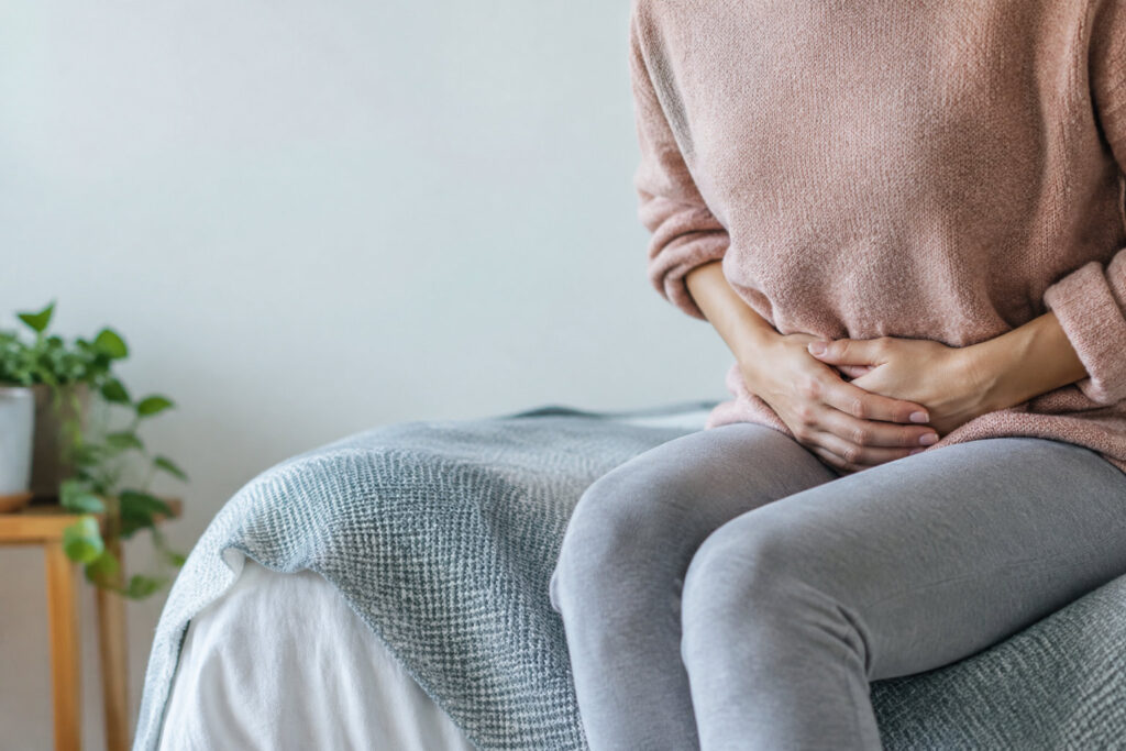 Une femme assise sur un lit, tenant son bas-ventre avec douleur, vêtue d'un pull rose et d'un pantalon gris.