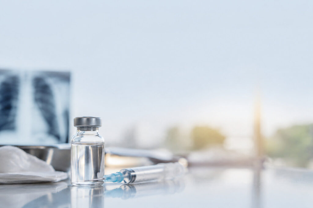 Fiole de vaccin et seringue posées sur une table médicale, avec une radiographie pulmonaire floue en arrière-plan.
