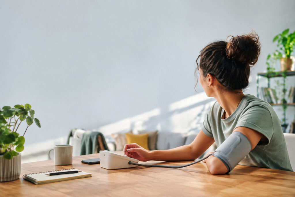 Une jeune femme assise à une table mesure sa tension artérielle avec un tensiomètre électronique.