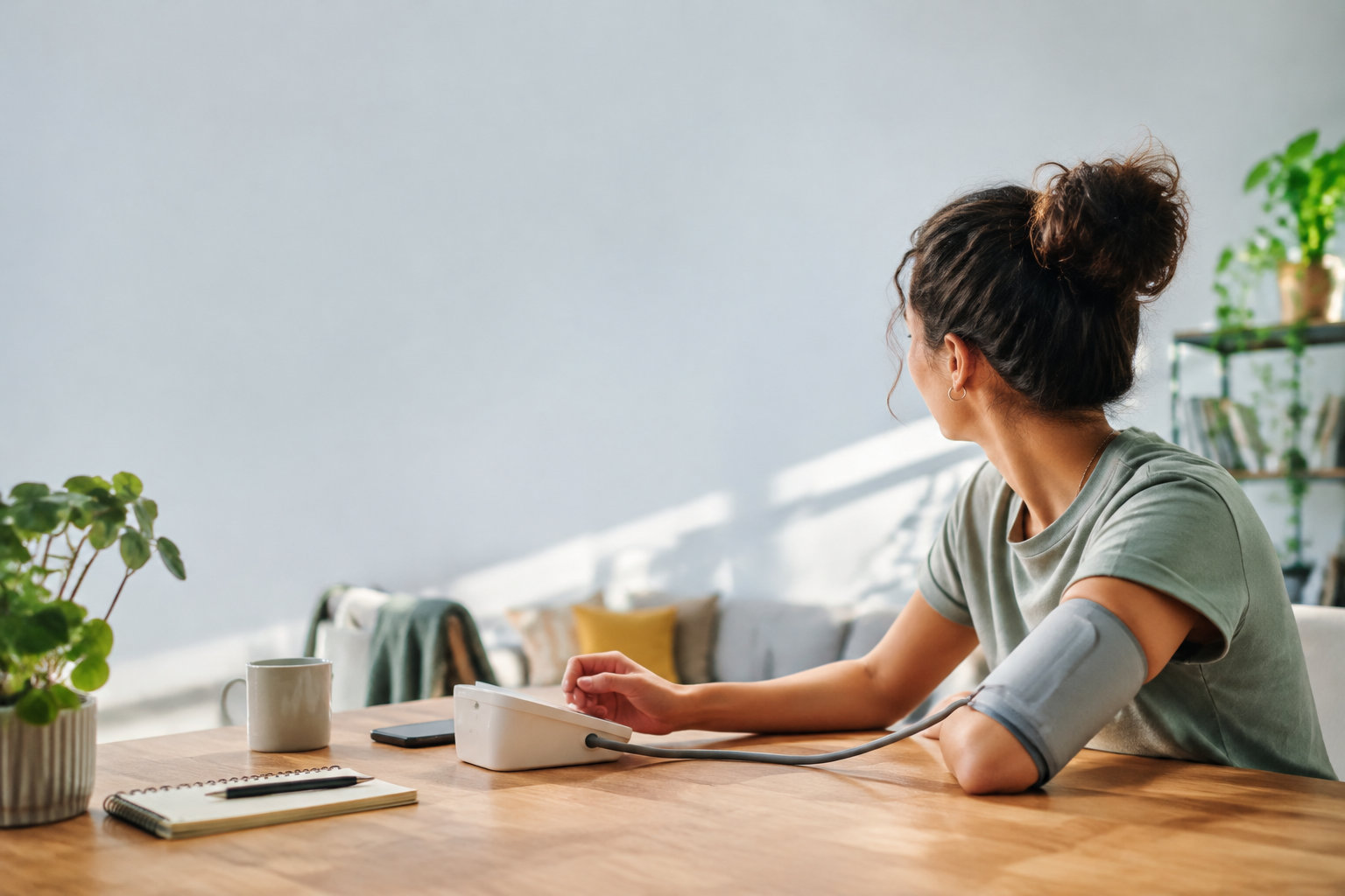 Une jeune femme assise à une table mesure sa tension artérielle avec un tensiomètre électronique.